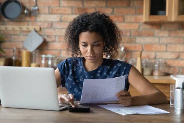 Serious millennial biracial woman calculate household expenditures finances on calculator at home. Focused African American wife pay bills taxes online on laptop, manage family finances or budget.