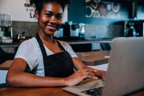 Barista a black woman in her coffee shop sits at counter with a laptop. Portrait of self-employed entrepreneur managing finance bookkeeping confidently in her own business.